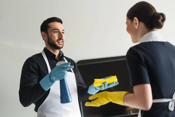 Cheerful housekeeper in rubber gloves talking with blurred chambermaid — Stock Photo