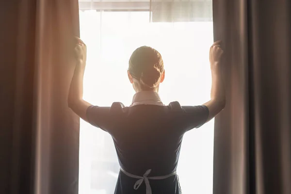 Vista trasera de la criada ajustando cortinas en la habitación de hotel - foto de stock