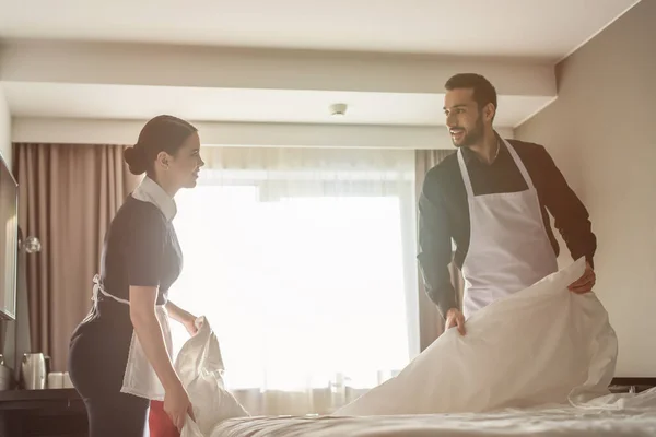 Donas de casa sorridentes olhando uns para os outros enquanto mudam de roupa de cama no quarto de hotel — Fotografia de Stock