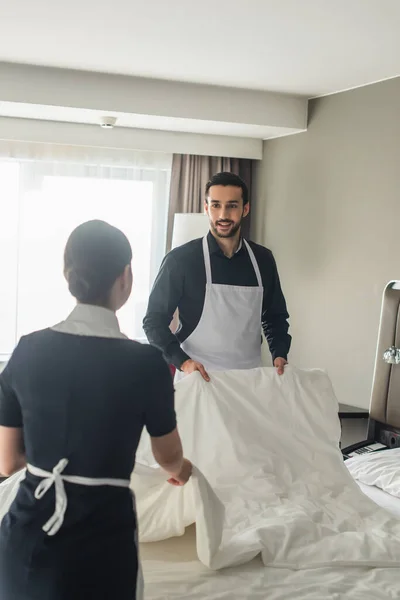 Lächelnd und bärtige Haushälterin wechselt Bettwäsche mit verschwommenem Dienstmädchen im Hotelzimmer — Stockfoto