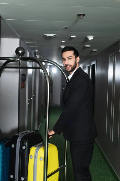 Fröhlicher Glockenspieler in der Nähe von metallischen Glockenwagen mit Gepäck in der Hotelhalle — Stockfoto