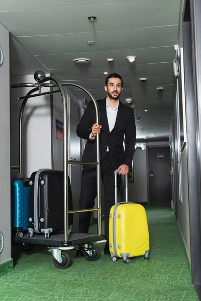 Pleine longueur de groom barbu près de chariot cloche métallique avec bagages dans le hall de l'hôtel — Photo de stock