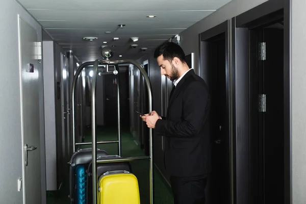 Bearded tourist messaging on smartphone near bell cart with luggage — Stock Photo