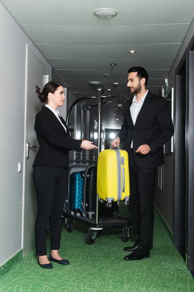 Lächelnde Empfangsdame zeigt mit der Hand in die Nähe des Kellners mit Gepäck — Stockfoto