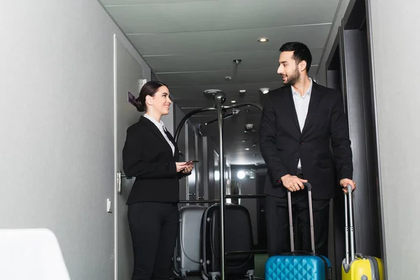 Receptionist sorridente che tiene smartphone e guarda fattorino con bagagli — Foto stock