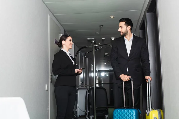 Lächelnde Empfangsdame mit Smartphone und Blick auf Kellner mit Gepäck — Stockfoto