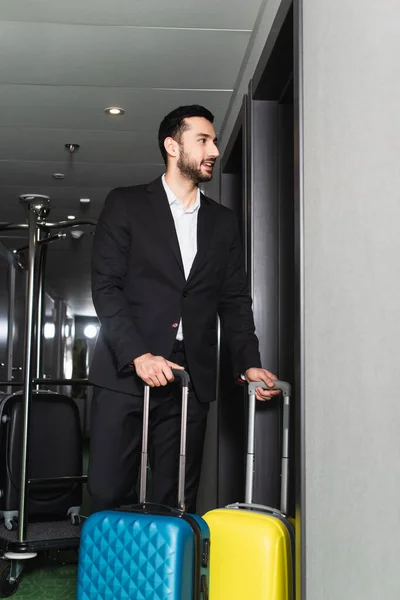 Hombre alegre y barbudo de pie con equipaje en el pasillo del hotel - foto de stock