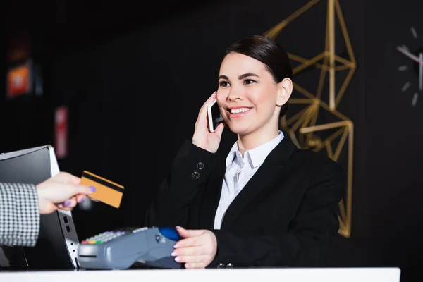 Recepcionista feliz sorrindo e falando no smartphone perto de convidado com cartão de crédito — Fotografia de Stock