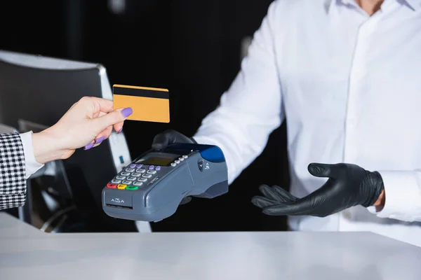 Vue recadrée de la réceptionniste en gants de latex tenant terminal de paiement près du touriste avec carte de crédit — Photo de stock