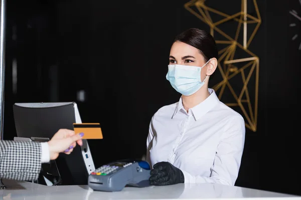Recepcionista en guantes de látex y máscara médica que sostiene la terminal de pago cerca de los huéspedes con tarjeta de crédito - foto de stock