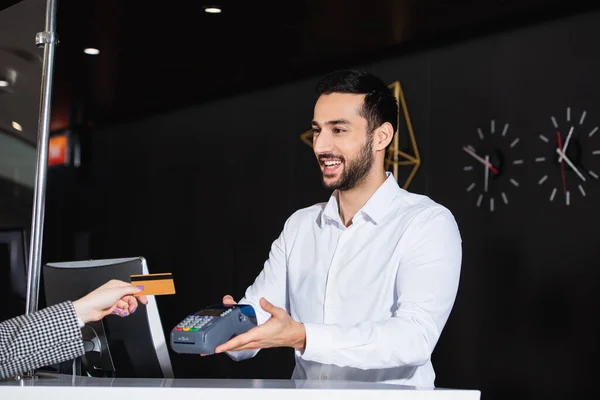 Réceptionniste barbu souriant tout en tenant terminal de paiement près du client avec carte de crédit — Photo de stock