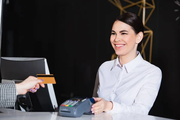 Feliz recepcionista sonriendo cerca de invitados pagando con tarjeta de crédito - foto de stock