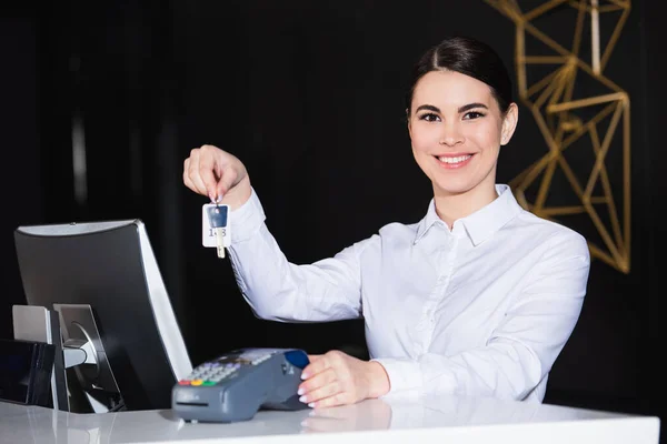 Recepcionista feliz segurando a chave do quarto perto do leitor de cartão de crédito no balcão — Fotografia de Stock