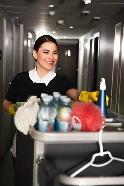 Cheerful maid in rubber gloves reaching spray bottle in housekeeping cart — Stock Photo