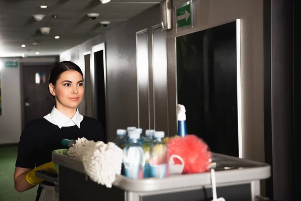 Young housekeeper in uniform near cart with cleaning supplies — Stock Photo