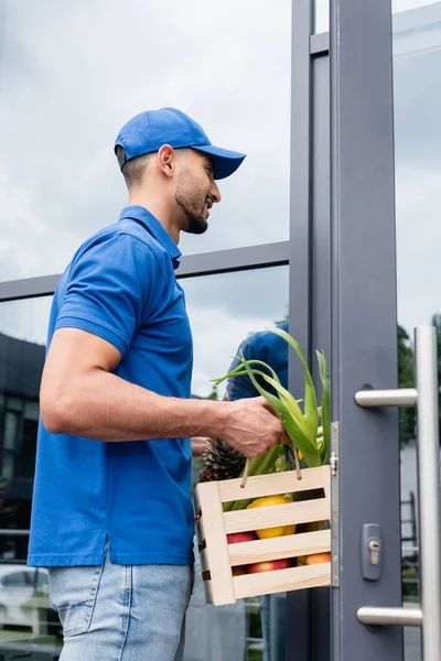 Smiling arabian courier holding box with fruits near door of building — Stock Photo
