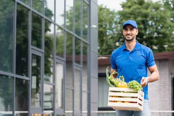 Positive arabian deliveryman holding box with vegetables near building — Stock Photo