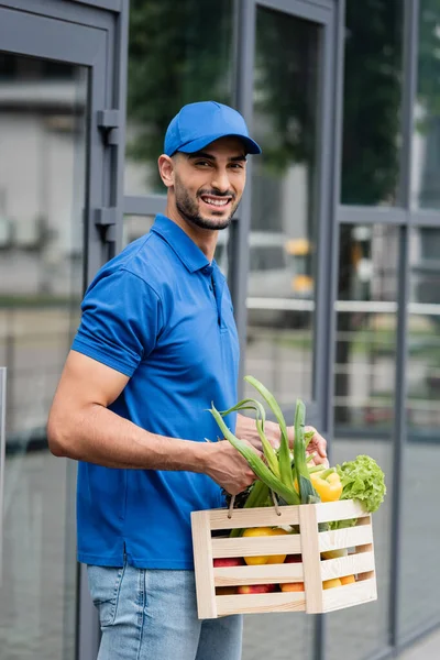 Arabian courier smiling at camera while holding box with vegetables outdoors — Stock Photo