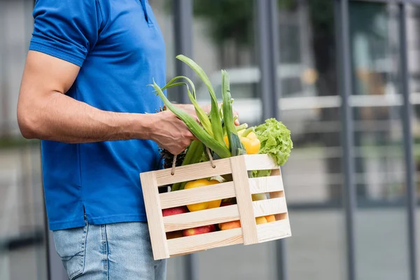 Cropped view of courier holding wooden box with vegetables outdoors — Stock Photo