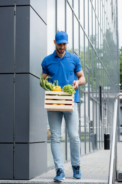 Correio árabe sorridente com legumes na caixa andando perto do edifício ao ar livre — Fotografia de Stock