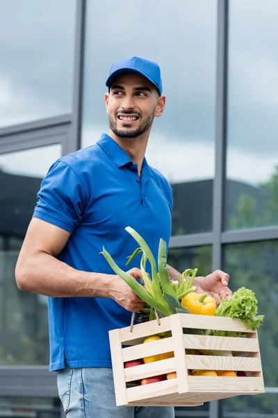 Happy arabian deliveryman holding box with fresh vegetables near building outdoors — Stock Photo