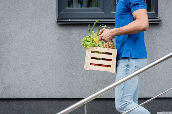 Vue recadrée du livreur tenant la boîte avec des légumes frais près du bâtiment — Photo de stock