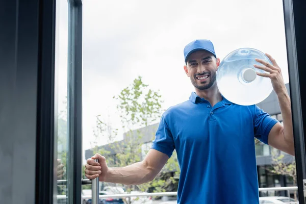 Alegre mensajero musulmán sosteniendo botella de agua cerca de la puerta del edificio - foto de stock