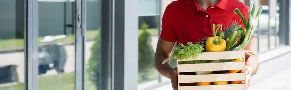 Vista cortada de jovem correio segurando caixa com vegetais orgânicos perto de construção, banner — Fotografia de Stock