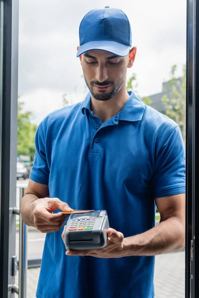 Arabian courier holding terminal de pago y tarjeta de crédito cerca de la puerta — Stock Photo