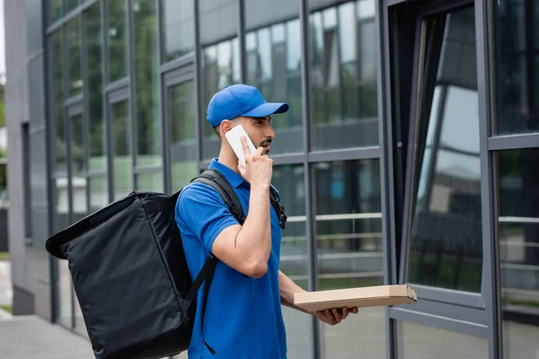 Vista lateral del mensajero árabe con caja de pizza hablando en el teléfono inteligente cerca del edificio - foto de stock