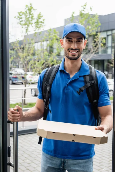 Alegre musulmán repartidor sosteniendo caja de pizza cerca de la puerta — Stock Photo