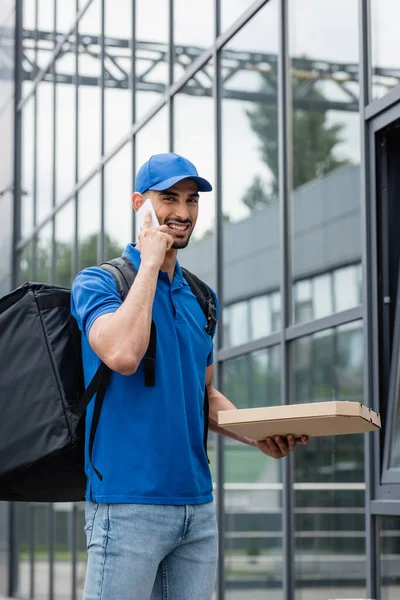 Smiling arabian deliveryman with backpack and pizza box talking on smartphone outdoors — Stock Photo