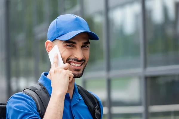 Mensajero árabe positivo con mochila hablando en el teléfono inteligente al aire libre - foto de stock