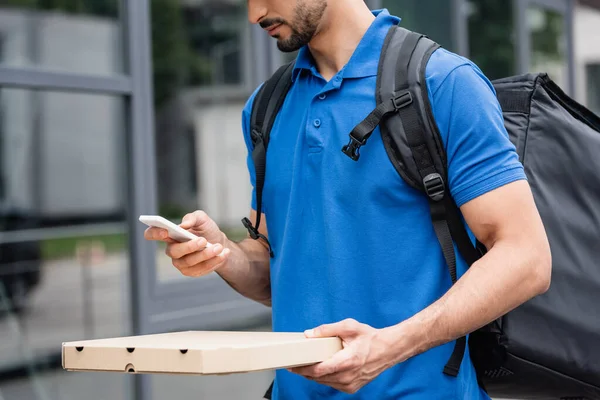 Vista recortada del repartidor sosteniendo la caja de pizza y el teléfono inteligente al aire libre - foto de stock