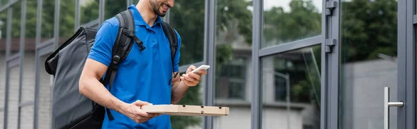 Vista recortada del mensajero sonriente sosteniendo el teléfono móvil y la caja de pizza cerca del edificio, pancarta - foto de stock