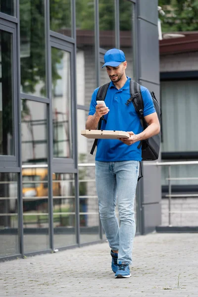 Cheerful arabian deliveryman using smartphone while holding pizza box near building — Stock Photo