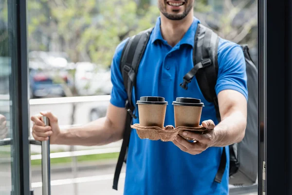 Cropped view of smiling courier on blurred background holding takeaway coffee near door — Stock Photo