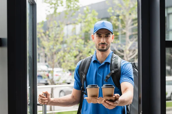 Mensajero árabe en uniforme sosteniendo café para ir cerca de la puerta - foto de stock