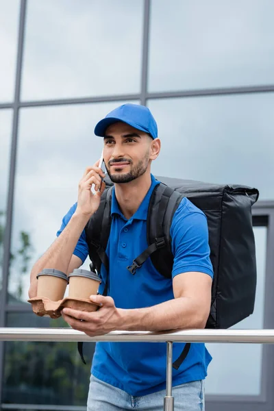 Young arabian courier with thermo backpack talking on smartphone and holding coffee to go near building outdoors — Stock Photo