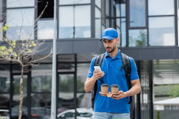 Mensajero árabe en uniforme usando celular y sosteniendo bebida para llevar en la calle urbana - foto de stock