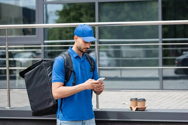 Giovane corriere musulmano con zaino termo utilizzando il telefono cellulare vicino al caffè per andare e costruire — Foto stock