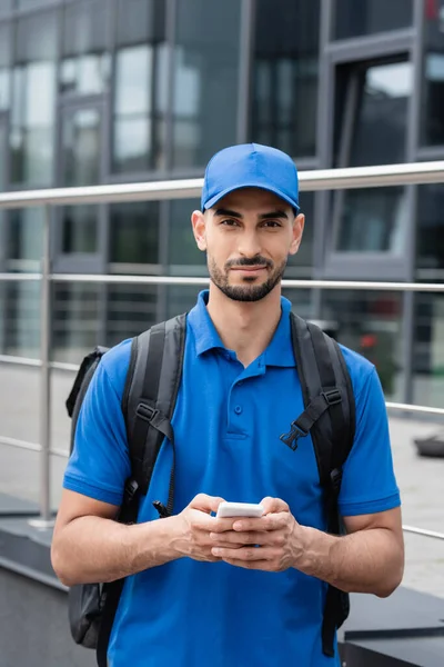 Jeune livreur musulman en uniforme et sac à dos tenant le téléphone portable et regardant la caméra à l'extérieur — Photo de stock