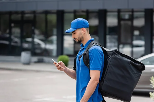 Vista lateral del mensajero musulmán con mochila térmica usando teléfono inteligente al aire libre - foto de stock