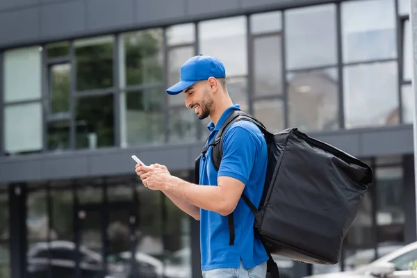 Side view of cheerful arabian courier with thermo backpack using smartphone on urban street — Stock Photo