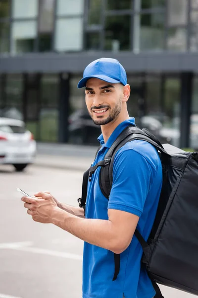 Jovem mensageiro árabe com mochila térmica e smartphone olhando para a câmera na rua urbana — Fotografia de Stock