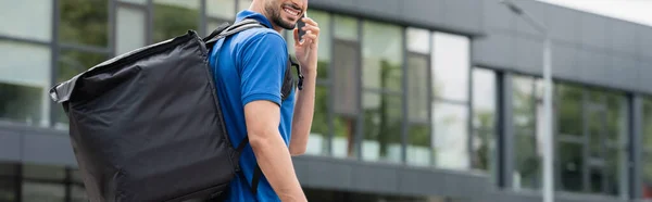 Vista recortada del repartidor sonriente con la mochila termo hablando en el teléfono inteligente al aire libre, bandera - foto de stock