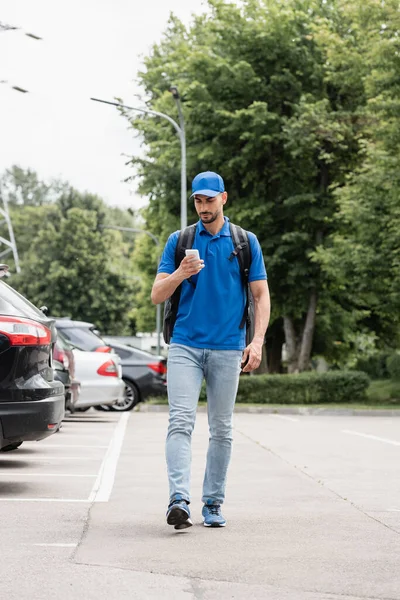 Joven repartidor árabe con mochila usando smartphone mientras camina por la calle urbana - foto de stock