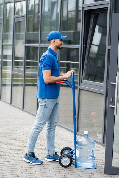 Vue latérale du courrier musulman souriant debout près de la bouteille d'eau sur le chariot et le bâtiment — Photo de stock