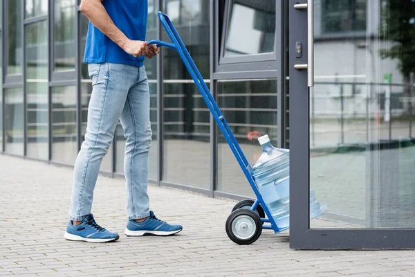 Ausgeschnittene Ansicht des Kuriers, der Karren mit Wasserflasche in der Nähe der offenen Tür des Gebäudes zieht — Stockfoto