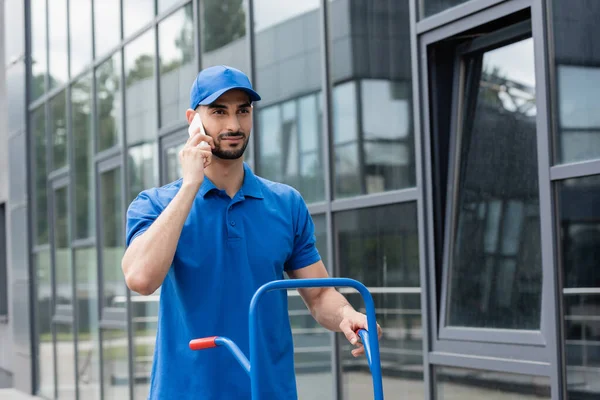 Joven repartidor musulmán hablando en el teléfono inteligente cerca del carro y el edificio al aire libre - foto de stock